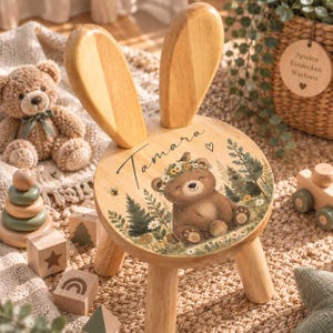 Peut inclure: Un tabouret rond en bois avec deux grandes oreilles de lapin dressées à l'arrière. L'assise présente une illustration imprimée d'un ours brun portant une couronne de fleurs, entouré d'arbres et de fleurs, avec le prénom Tamara et un cœur.