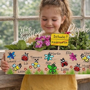 Könnte beinhalten: Ein hölzerner Pflanzkasten mit einem Design aus Insekten und Blumen, gehalten von einer Person. Die Box trägt Namen und die Wörter "Schule" und "Kindergarten" auf einem Schild. Die Box ist mit Pflanzen und Blumen gefüllt.