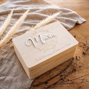 Peut inclure: Un coffret souvenir en bois clair avec le nom "Marie" en écriture blanche. La boîte présente un motif de couronne florale et du texte gravé. La boîte est posée sur une surface en bois avec des éléments décoratifs.
