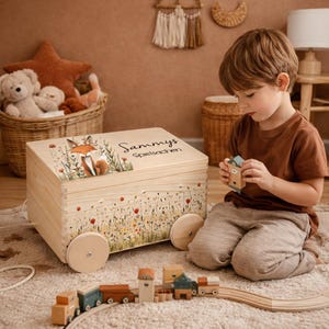 Può includere: Un baule di giocattoli in legno con un'illustrazione di volpe e la scritta "Sammys Spielsachen". Il baule ha un design floreale ed è su ruote. Un bambino gioca con una casetta di legno e un trenino di legno.