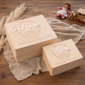 May include: Two light-coloured wooden keepsake boxes, one large and one small, with the name "Marie" in cursive script. The boxes have engraved details including a date, weight, and height. The boxes are on a neutral-coloured fabric.