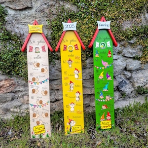 Może przedstawiać: Trzy drewniane tablice wzrostu z domem na szczycie i czerwonym, żółtym i zielonym tłem. Każda tablica ma inny motyw zwierzęcy i zawiera imiona i liczby. Tekst na tablicach brzmi "Wer ziehen wetter" i "Schule 1 Kindergarten".