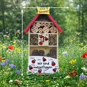 May include: A wooden insect house, 60cm tall and 30cm wide, with a red roof and multiple compartments filled with bamboo and drilled wood. The sign reads "Wir sind ausgeflogen" with names. Includes a yellow sign that says "Schule &uarr; Kindergarten".