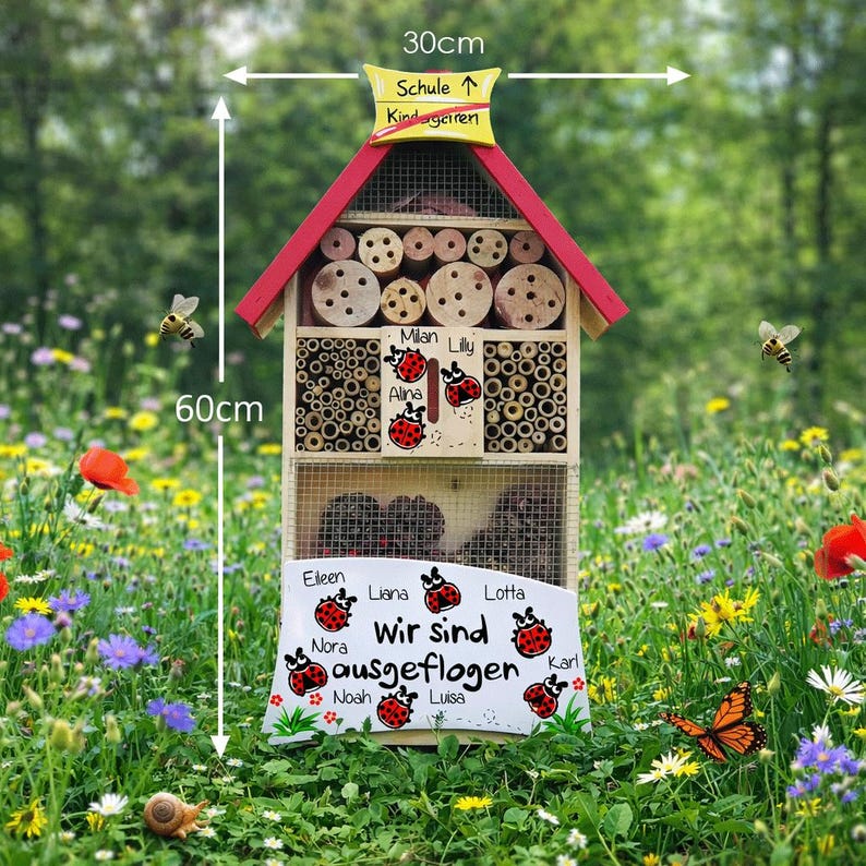 May include: A wooden insect house, 60cm tall and 30cm wide, with a red roof and multiple compartments filled with bamboo and drilled wood. The sign reads "Wir sind ausgeflogen" with names. Includes a yellow sign that says "Schule &uarr; Kindergarten".