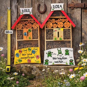 May include: Two wooden insect houses with red roofs, labelled 2022 and 2024. The houses are filled with bamboo and wooden discs. A yellow measuring tape shows 60 cm. Decorative signs with text and frog illustrations are below the houses.