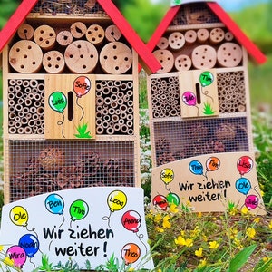 May include: Wooden insect houses with red roofs, featuring various compartments filled with bamboo and wood. The houses have signs with names and the phrase "Wir ziehen weiter!" written on them. The houses are set in a grassy area.