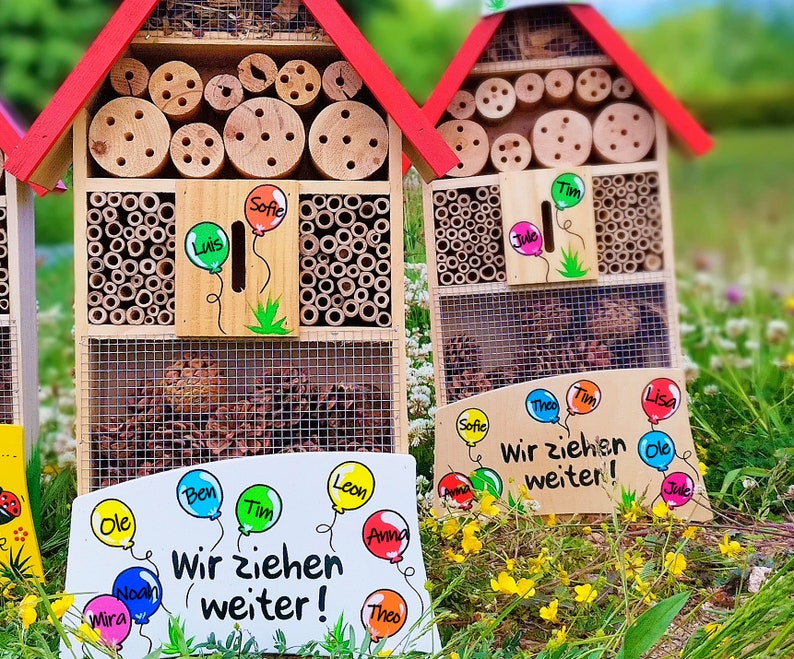 May include: Wooden insect houses with red roofs, featuring various compartments filled with bamboo and wood. The houses have signs with names and the phrase "Wir ziehen weiter!" written on them. The houses are set in a grassy area.