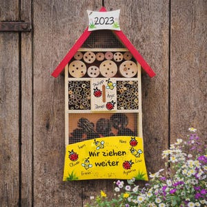 May include: A wooden insect hotel with a red roof and a sign that reads "2023". The hotel has multiple compartments filled with bamboo sticks and wooden discs. A yellow sign displays the text "Wir ziehen weiter" and names.