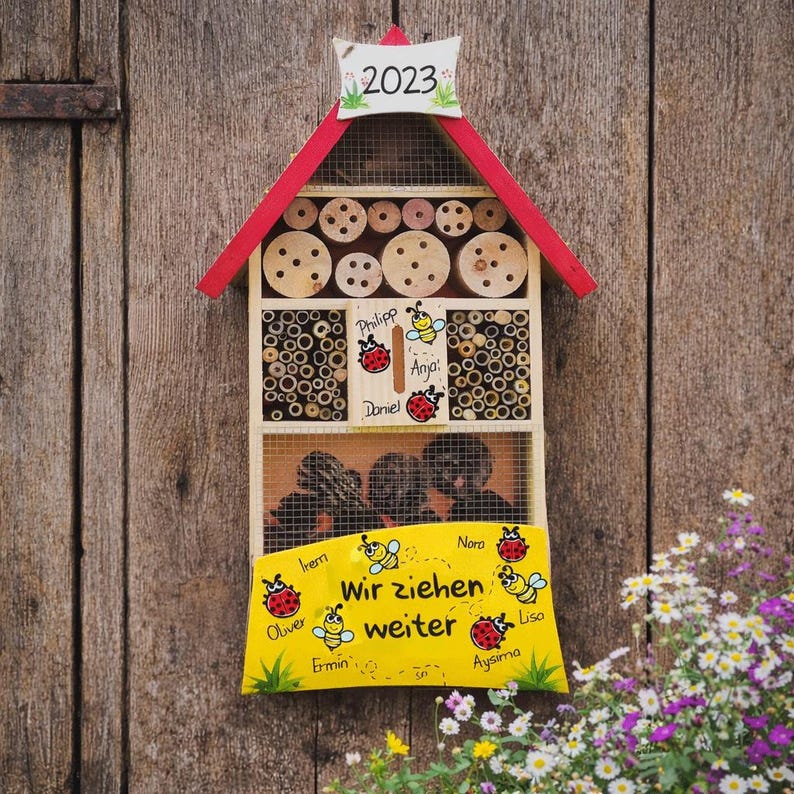 May include: A wooden insect hotel with a red roof and a sign that reads "2023". The hotel has multiple compartments filled with bamboo sticks and wooden discs. A yellow sign displays the text "Wir ziehen weiter" and names.