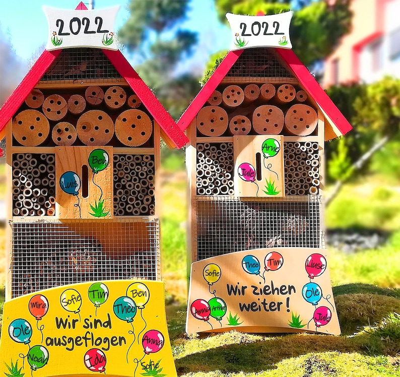 May include: Two wooden insect houses with red roofs and the year "2022" on top. The houses have various openings filled with bamboo and wood. Each house has a sign with names and balloon illustrations. One sign reads "Wir sind ausgeflogen" and the other reads "Wir ziehen weiter!"