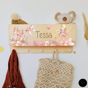Peut inclure: Un porte-manteau en bois avec un motif de sirène et de tortue de mer rose et blanc. Le porte-manteau a trois crochets et le nom "Tessa" est écrit sur le devant.