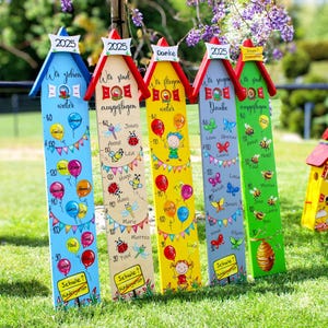 Może przedstawiać: Pięć kolorowych drewnianych tablic wzrostu z daszkami w kształcie domów, każda w innym kolorze i wzorze. Tablice mają miary, imiona i elementy dekoracyjne, takie jak balony i owady. Tekst zawiera rok 2025 i słowo "Kindergarten".