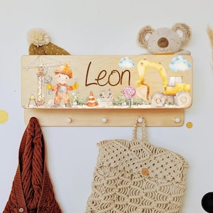 Peut inclure: Porte-manteau mural en bois avec le nom "Leon" et des illustrations sur le thème de la construction. Comprend une grue, un ouvrier, une excavatrice et un panneau stop. Avec des chevilles pour suspendre des articles. Un jouet en peluche koala est posé dessus.