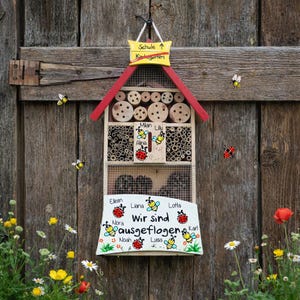 May include: A wooden insect hotel with a red roof and a sign that says "Schule Kindergarten". The hotel is decorated with bees, ladybirds, and the words "Wir sind ausgeflogen". The hotel is hanging on a wooden wall.