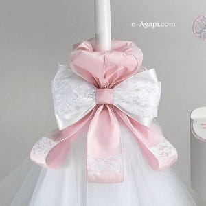 Può includere: Una candela battesimale in tulle bianco e raso rosa con un grande fiocco rosa e un bordo in pizzo bianco. La candela è decorata con un nastro rosa e un fiocco in pizzo bianco. La candela è su uno sfondo bianco.
