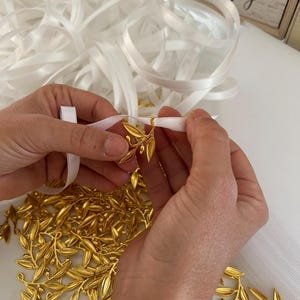 May include: A pile of gold-colored olive branch charms with white satin ribbon. The ribbon is tied around the charms.