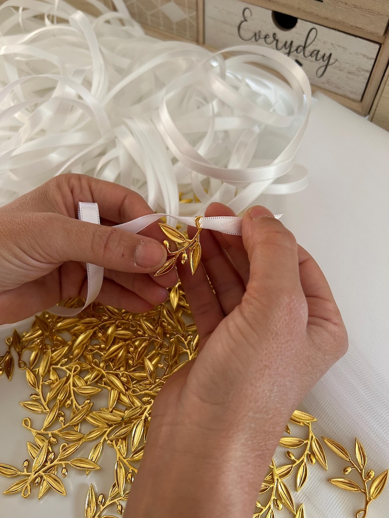 May include: A pile of gold-colored olive branch charms with white satin ribbon. The ribbon is tied around the charms.