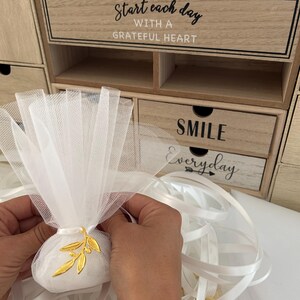 May include: White tulle bags with gold olive branch charms, white satin ribbon, and a wooden box with the text "Start each day with a grateful heart" and "Smile everyday".
