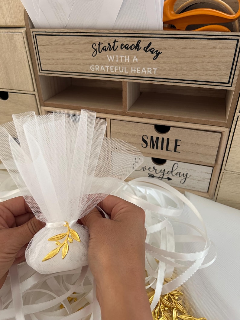 May include: White tulle bags with gold olive branch charms, white satin ribbon, and a wooden box with the text "Start each day with a grateful heart" and "Smile everyday".