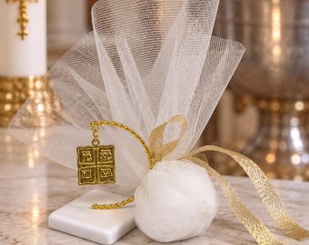 May include: A white tulle favor bag tied with a gold ribbon, featuring a gold-toned square charm embossed with the Greek Orthodox IC XC NI KA inscription. The charm hangs from a curved gold wire stand attached to a small white rectangular base.