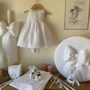 May include: White christening gown with a white satin bow, a white candle with a white satin bow, a white bonnet, a pair of white baby shoes, a white blanket, a white box with a white satin bow, and a small glass bottle with a white ribbon.