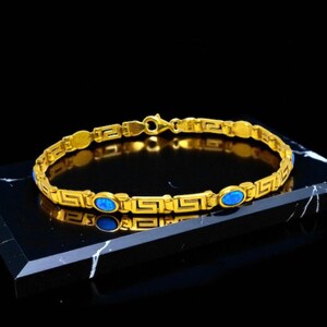 Op de afbeelding: Een goudkleurige armband met een Grieks sleutelmotief en ovale blauwe edelstenen. De armband, met een sluiting, is gemaakt van glanzend metaal. Het wordt gepresenteerd op een zwart-witte ondergrond. Het ontwerp van de armband is ingewikkeld.