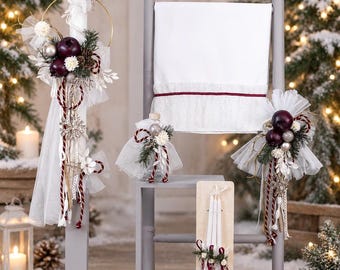 May include: A set of white candles and a white cloth featuring decorative arrangements of dark red ornaments, pine sprigs, white tulle, and braided ribbons. The items are displayed on gray stands and a wooden board, accented with small white flowers and silver details.