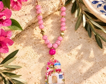 May include: A beaded necklace featuring pink marbled beads, small gold-toned cross spacers, and a central pendant shaped like a building with a blue dome, pink flowers, and a gold cross. The necklace is strung on a thin gold-toned wire.