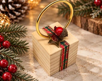 May include: A square wooden block ornament featuring a gold-toned metal ring handle, wrapped with a red and green plaid ribbon. The decoration is accented with a small gold star and a single red berry attached to a textured brown acorn cap.