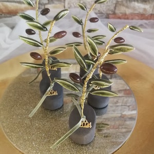 May include: Four small olive branch sculptures with brown olives and green leaves. Each sculpture is on a black base with a gold 2024 charm. The sculptures are on a round, gold glitter surface.