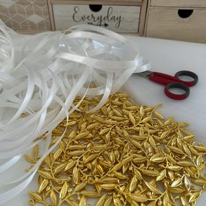 May include: A pile of gold-colored metal olive branch charms with a white satin ribbon and red scissors on a white surface. The background includes a wooden box with the text "SMILE Everyday".