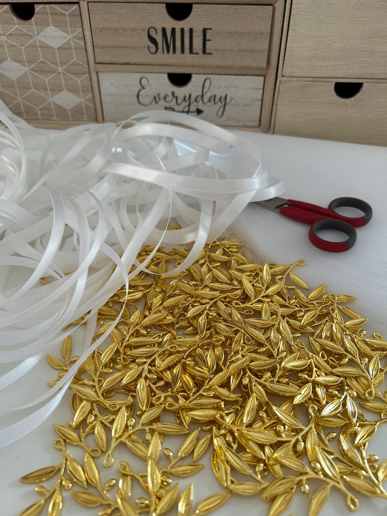 May include: A pile of gold-colored metal olive branch charms with a white satin ribbon and red scissors on a white surface. The background includes a wooden box with the text "SMILE Everyday".