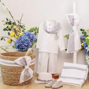 Puede incluir: Un conjunto de lino blanco para un bebé, que incluye una camisa, pantalones, sombrero y una bufanda con rayas grises y blancas. El conjunto se muestra con un lazo blanco y gris en una cesta de mimbre, un lazo blanco y gris en una toalla blanca y un lazo blanco y gris en una vela blanca.