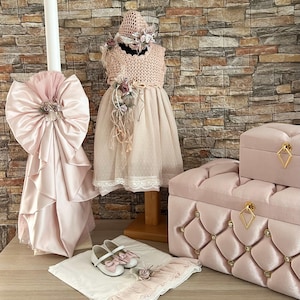 Puede incluir: Un conjunto de bautizo rosa para bebé con un vestido de ganchillo, un sombrero, un lazo de satén, un par de zapatos blancos, una manta blanca con ribete de encaje y dos cajas de terciopelo rosa con detalles dorados.