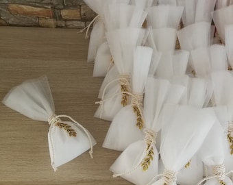 May include: A group of white tulle bags with gold leaf accents. The bags are tied with a white ribbon and are arranged in a pyramid shape. The bags are on a wooden surface.