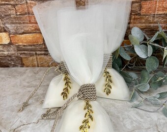 May include: Three white tulle favor bags tied with brown rope and gold leaf charms. The bags are filled with a white substance and are arranged on a marble surface. The background features a brick wall and greenery.