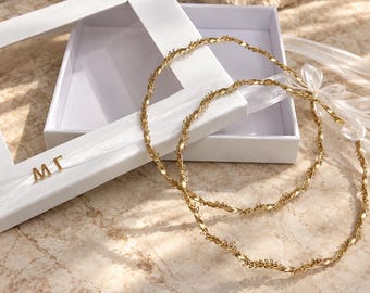 May include: Two circular gold-toned crowns featuring a twisted metal design with delicate leaf-like detailing. The crowns are connected by a white ribbon with the gold letters M and Γ attached, presented with a white rectangular box.
