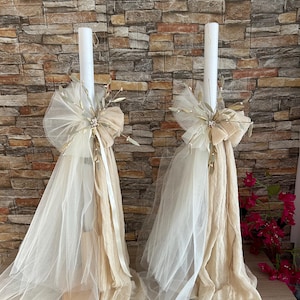 May include: Two white candles with beige tulle and gold accents. The candles are decorated with sprigs of greenery and a bow. The candles are on wooden bases.