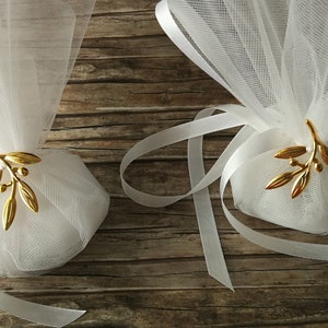 May include: Two white tulle favor bags with gold olive branch embellishments and white satin ribbon ties. The bags are on a wooden surface.