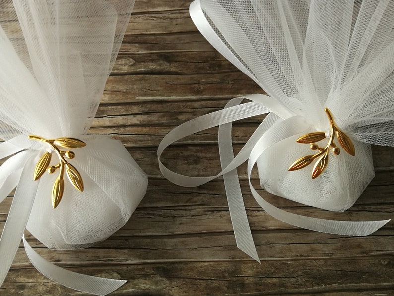 May include: Two white tulle favor bags with gold olive branch embellishments and white satin ribbon ties. The bags are on a wooden surface.