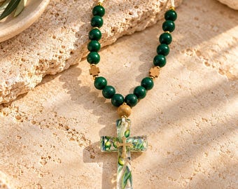 May include: A beaded necklace featuring dark green spherical beads, small wooden cross spacers, and a central clear resin cross pendant containing embedded green olive leaves and a small gold-toned cross, all strung on a light-colored cord.