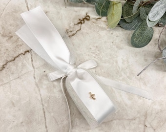 May include: A white ribbon tied in a bow, with a small gold-colored charm attached. The ribbon is wrapped around a small, rectangular object, possibly a gift or favor. The background features greenery and a marble-like surface.