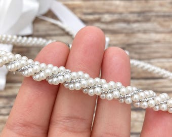 May include: A decorative trim featuring a twisted design of small white faux pearls intertwined with a shimmering silver metallic thread. The flexible strand is arranged in a continuous pattern, showcasing a textured combination of pearl and silver.