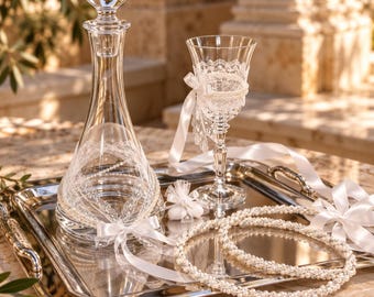 May include: A clear glass decanter with a spherical stopper, a matching stemmed glass, and two circular beaded wreaths on a silver rectangular tray. The decanter and glass are adorned with white lace trim, pearl accents, and white satin ribbons.