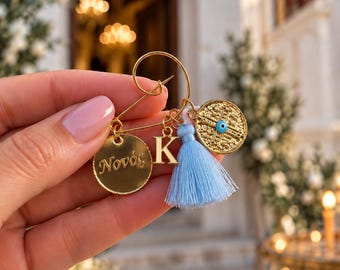 May include: A gold-toned safety pin brooch featuring a round charm with the Greek text Novós, a gold letter K charm, a light blue tassel, and a circular charm with an embossed pattern and a central blue evil eye bead accent.