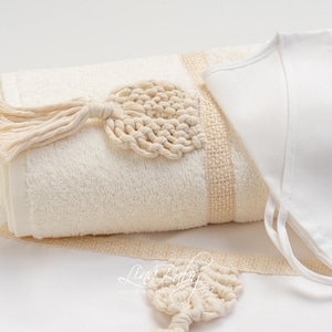 May include: A folded ivory towel with a woven beige stripe and a macrame leaf accent. A matching bib with tie closures and a macrame leaf are also visible. The items are arranged on a white surface.