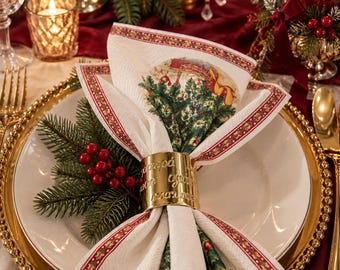 May include: A white napkin with a festive Christmas tree and sleigh print, featuring a red and green border, held by a polished gold-tone metal napkin ring engraved with Greek text, resting on a white plate with a sprig of green pine and red berries.