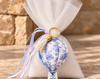 May include: A white tulle favor bag tied with light blue and pink ribbons, featuring a gold-toned keyring with a small white tassel and a padded fabric hot air balloon ornament decorated with a blue and white floral pattern.
