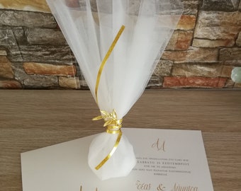 May include: A white tulle favor bag tied with a gold ribbon and a gold olive branch charm, placed on top of two cream-colored wedding invitations featuring gold lettering and the initials AA with the date 16.09.2023.