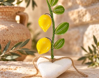 May include: A decorative sculpture featuring two bright yellow glass lemons attached to a green wire stem with several glossy green ceramic leaves. The stem is anchored into a smooth, white, heart-shaped stone base tied with a thin brown twine.